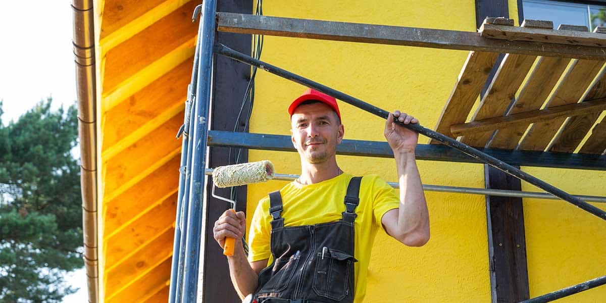 A house painter holds a roller near a bright yellow exterior wall, showing how temperature and humidity affect Las Cruces paint jobs.