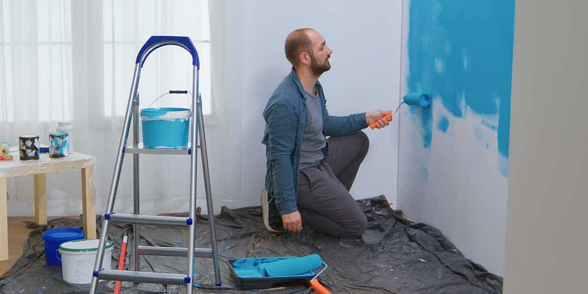 A man kneeling inside a room uses a paint roller to apply bright blue paint onto a white wall.